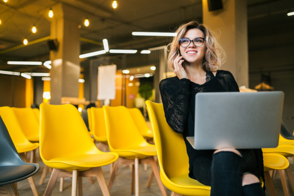 young attractive woman sitting lecture hall working laptop wearing glasses many yellow chairs student education online freelancer smiling talking smartphone looking forward startup