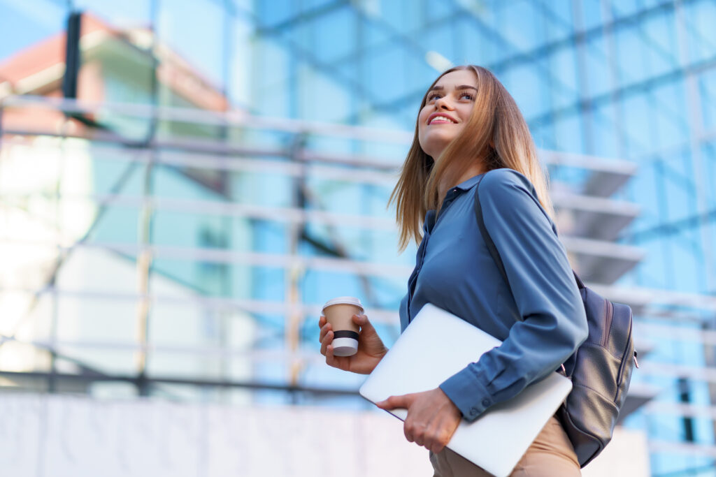 É o fim da carreira aspiracional? Por que as novas gerações não querem mais a vida dos seus líderes 1 blonde young woman smiling portrait holding laptop coffee wearing blue gentle shirt modern building