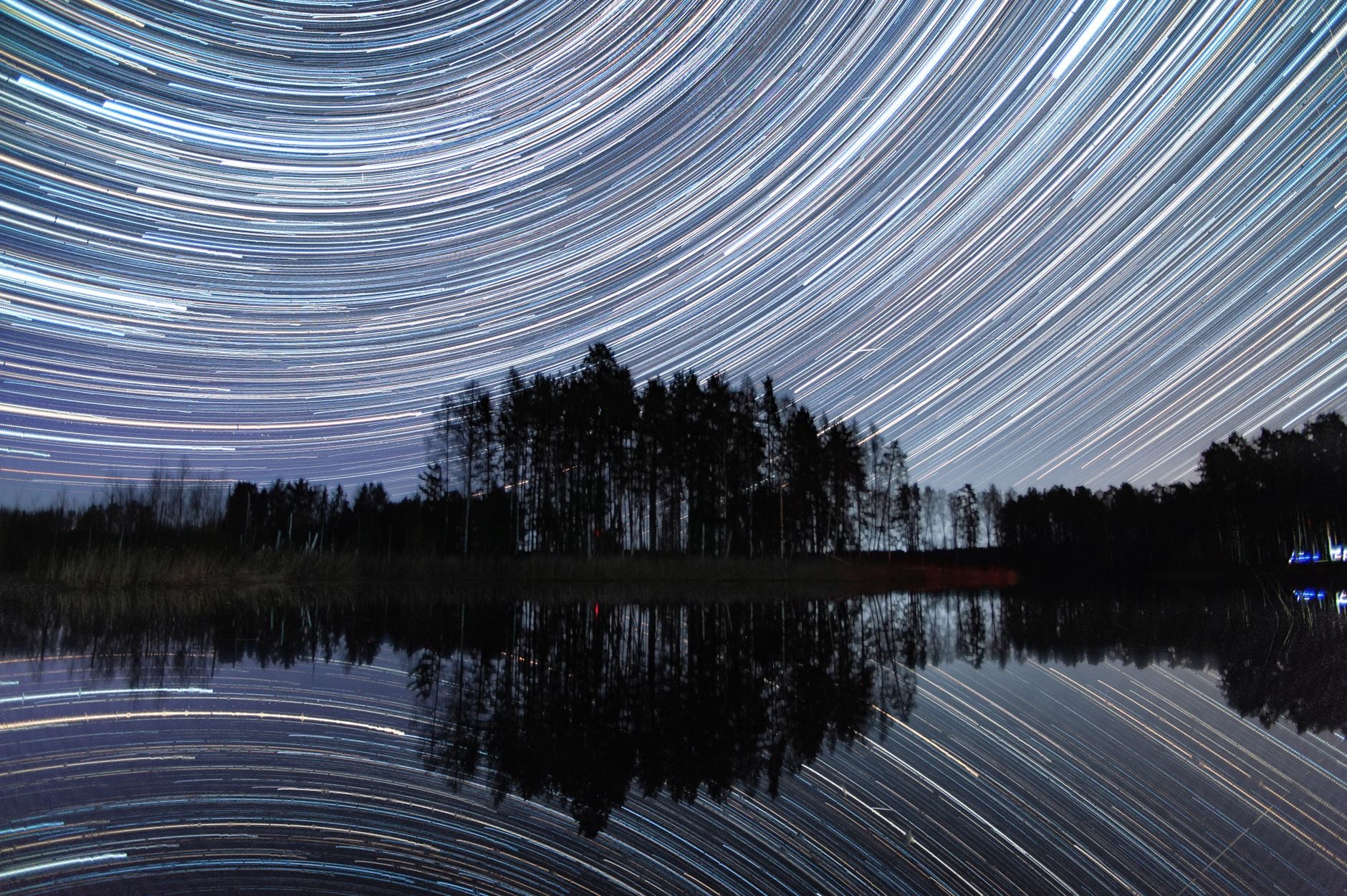 Trilhas de estrelas vívidas sobre um lago tranquilo com árvores refletindo na água, capturadas em uma foto em time-lapse.