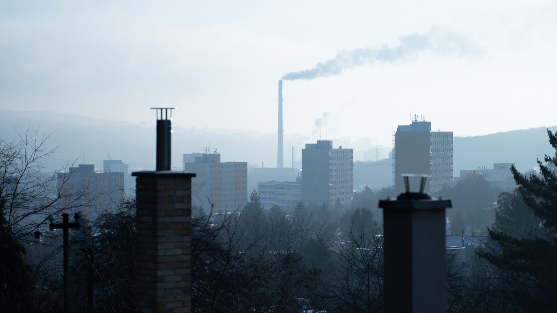 Vista de uma paisagem urbana com chaminés, fumaça e edifícios altos em um dia nublado.