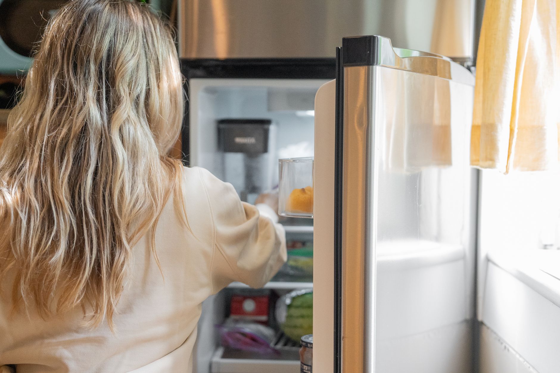 Nem tudo é simplesmente colocar no freezer: especialista explica como congelar alimentos sem comprometer sabor, textura e qualidade 1 Vista de costas de uma mulher loira retirando comida da geladeira em uma cozinha iluminada.