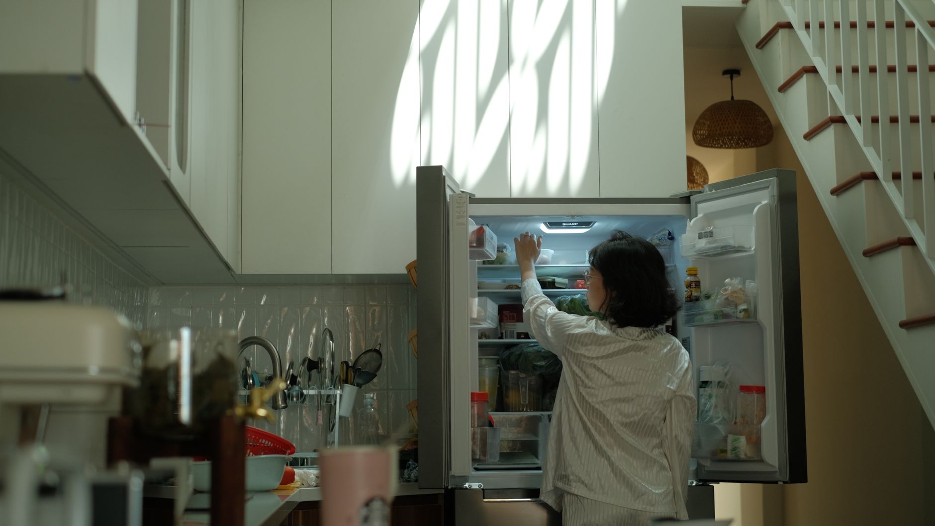 Nem tudo é simplesmente colocar no freezer: especialista explica como congelar alimentos sem comprometer sabor, textura e qualidade 2 Uma mulher de pijama verifica a geladeira em uma cozinha aconchegante e bem iluminada.