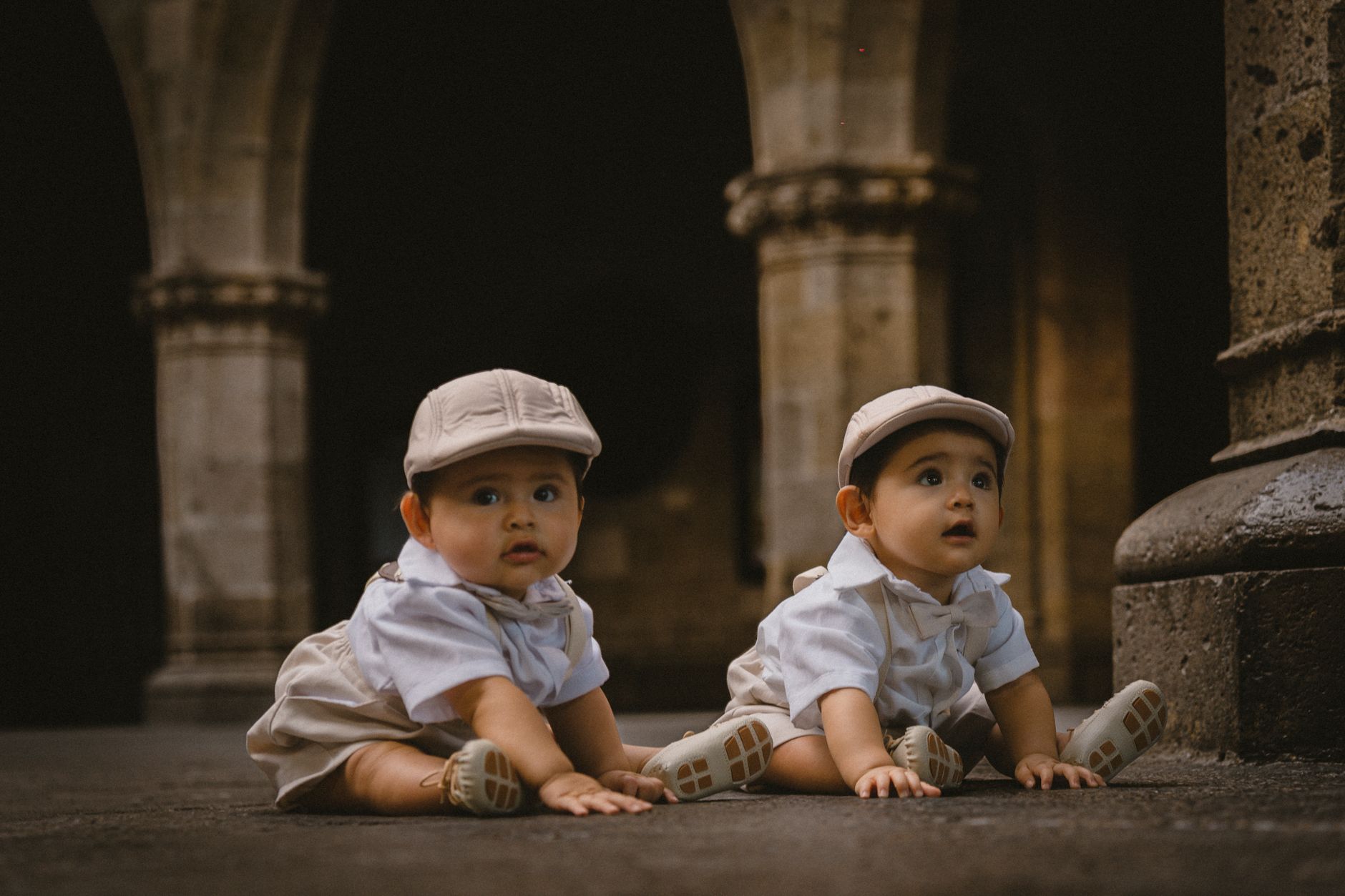 Pais brasileiros estão buscando nomes cada vez mais diferentes para os filhos, e três tendências que envolvem luxo, internet e futebol devem ganhar força em 2027 1 Adoráveis gêmeos bebês de boné sentados em uma antiga calçada de pedra com arcos ao fundo.