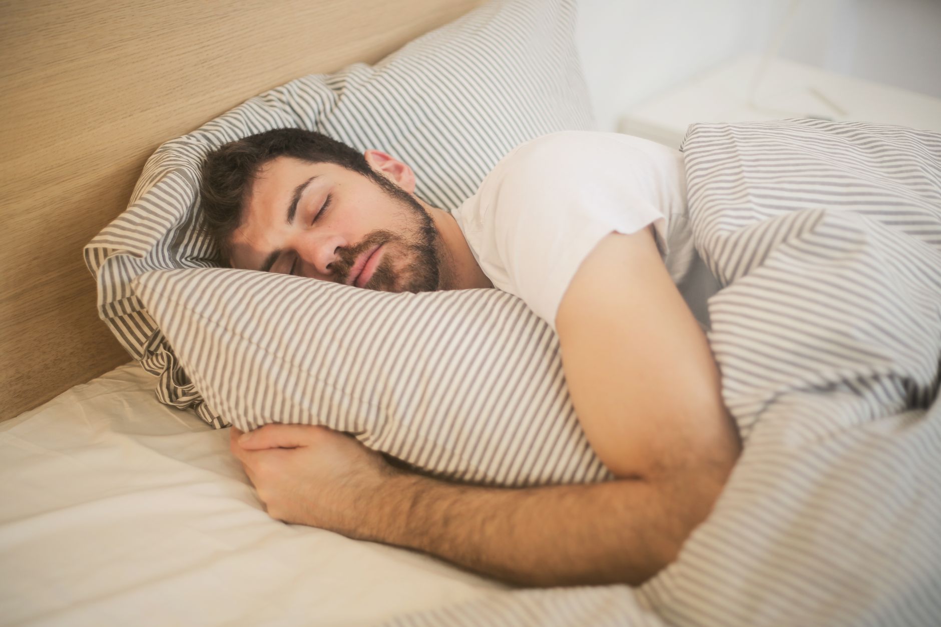 Por que algumas pessoas conversam, respondem e até gritam enquanto estão dormindo? Especialista explica o que pode estar por trás do comportamento 2 Homem dormindo pacificamente em roupas de cama listradas, abraçando relaxamento e conforto.