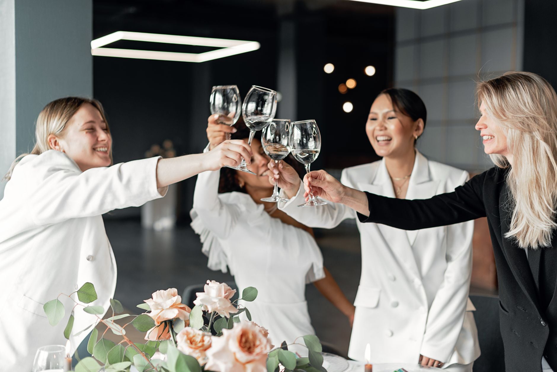 Mulheres celebrando em ambientes internos com um brinde, batendo taças de vinho, alegres e sorridentes.