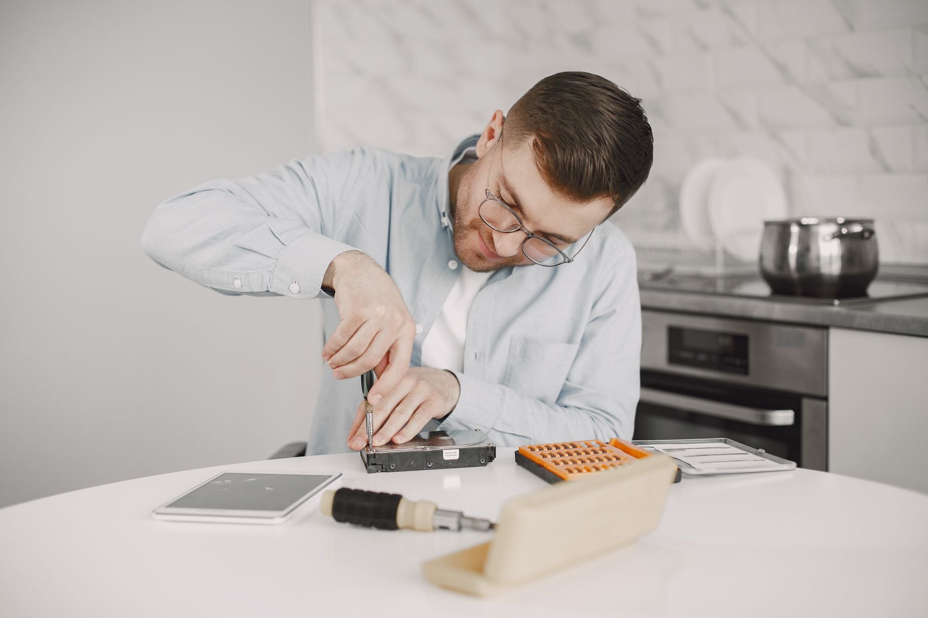 Homem usando óculos, utilizando uma chave de fenda para consertar um dispositivo em uma mesa de cozinha.
