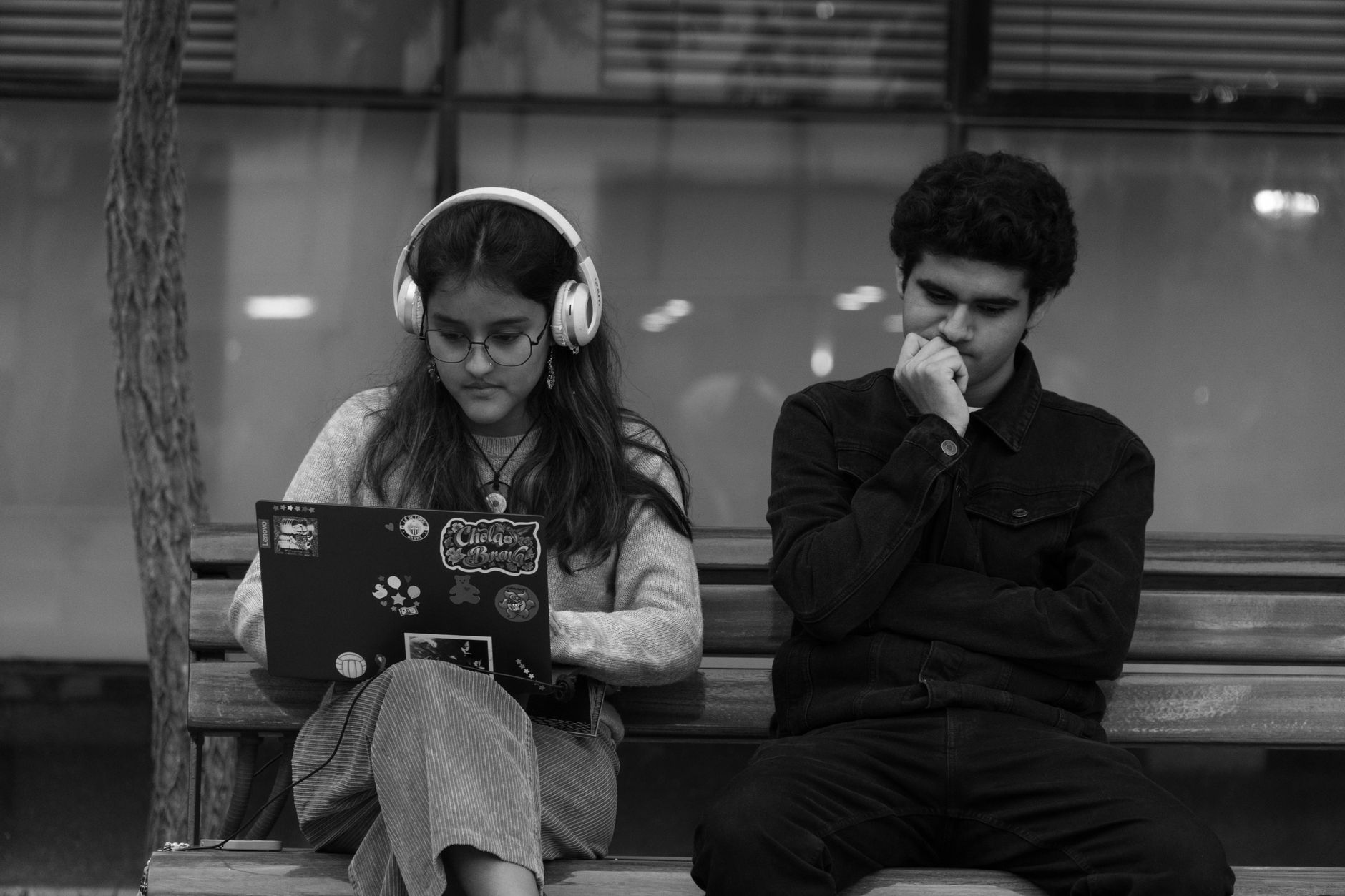 Imagem em preto e branco de adolescentes sentados em um banco, um com fones de ouvido e um laptop, o outro pensativo.