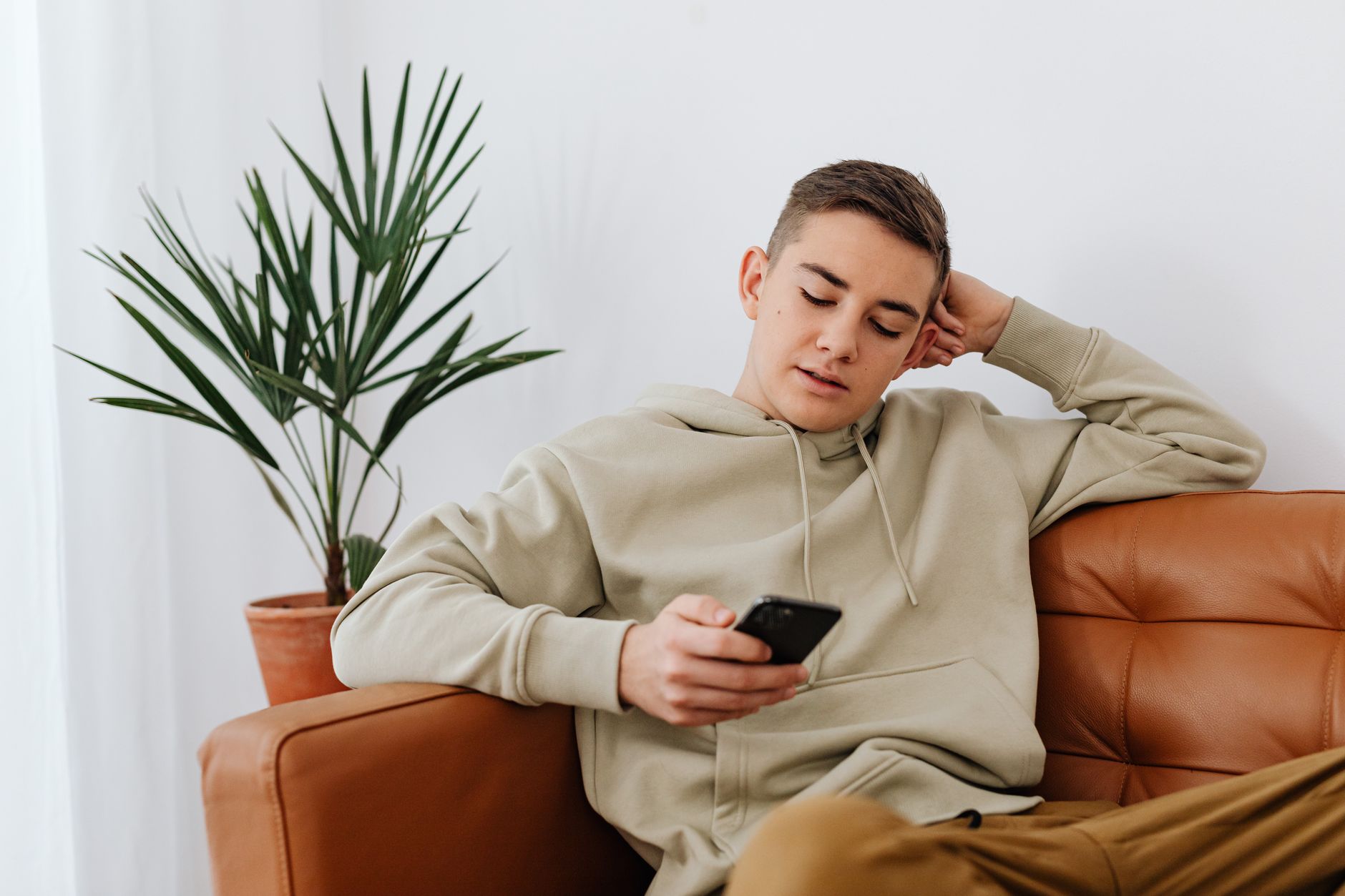 Jovem sentado em casa enviando mensagens em um smartphone, usando um moletom.