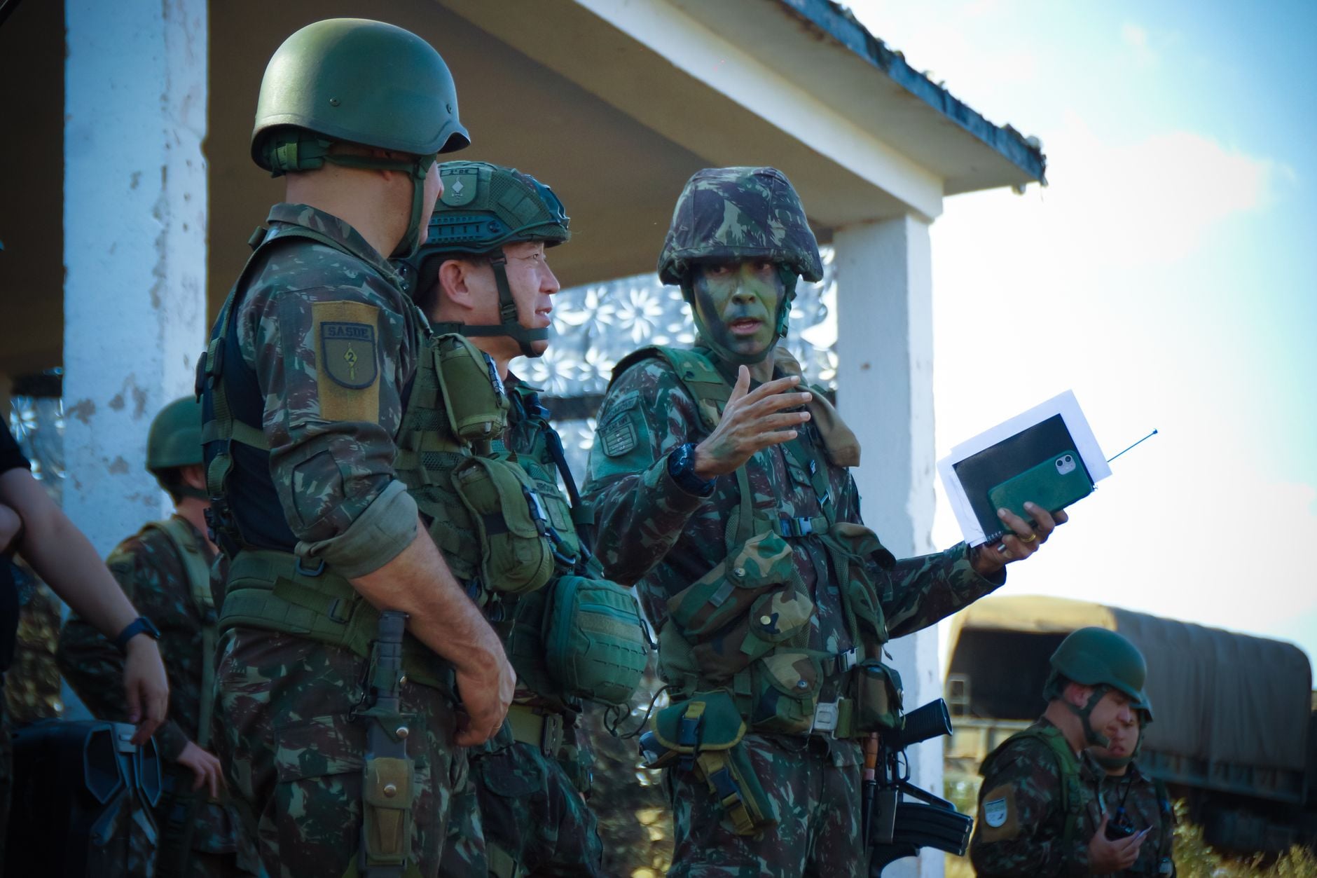 Grupo de soldados camuflados participando de uma reunião ao ar livre com equipamentos táticos e capacetes.