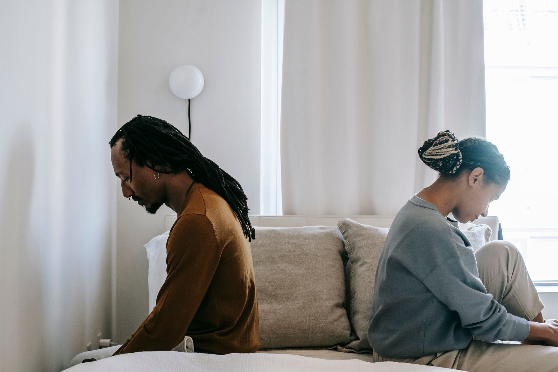 Casal afro-americano sentado de costas um para o outro em uma cama, retratando conflito de relacionamento em casa.