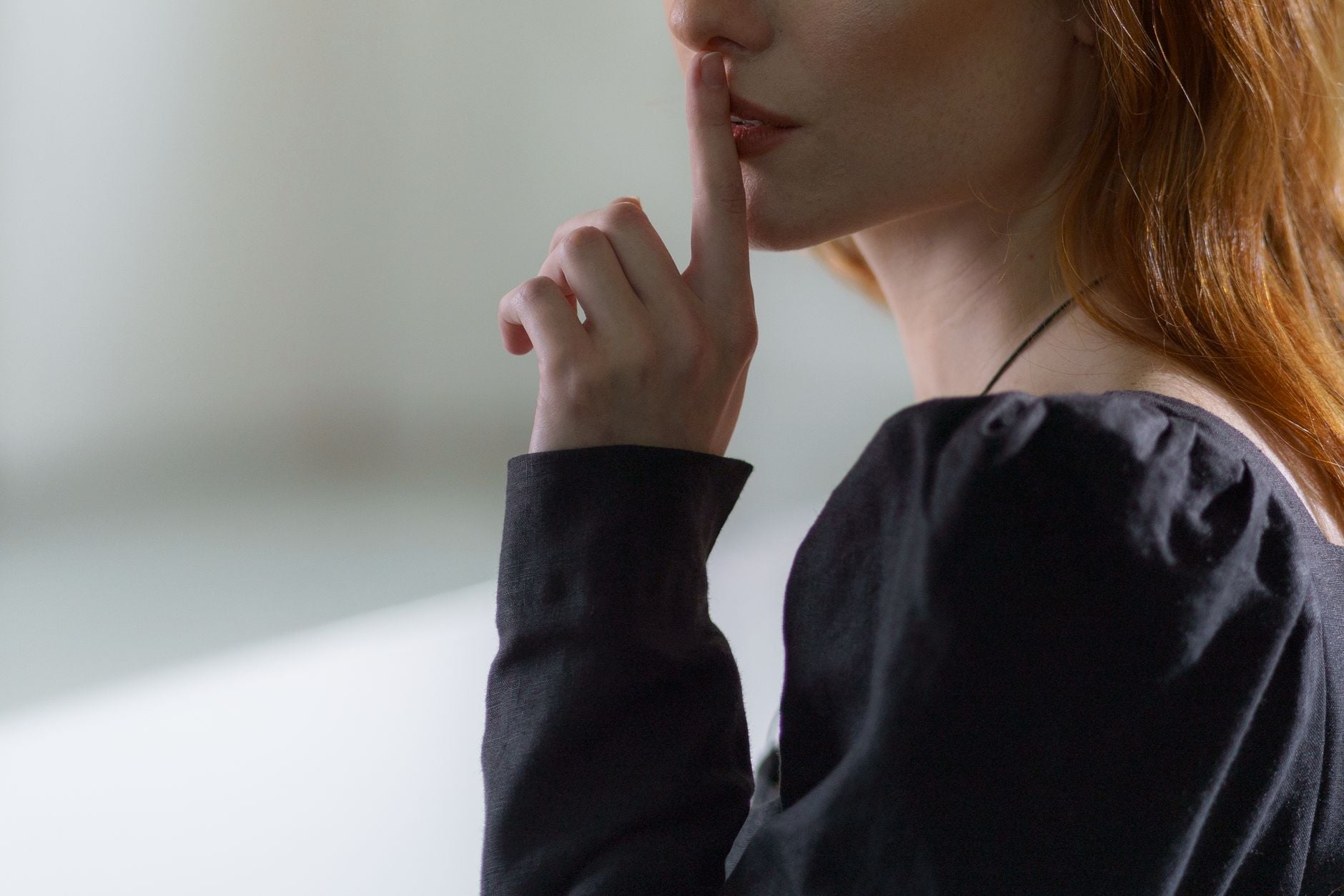 Close-up de mulher sugerindo silêncio com o dedo nos lábios, vestindo uma blusa preta.