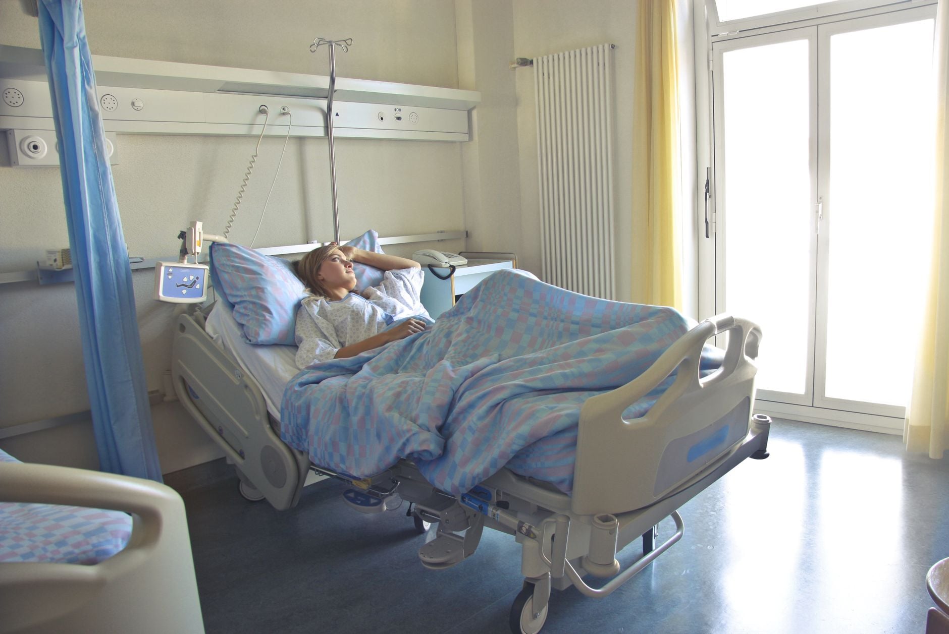 Uma mulher deitada em uma cama de hospital em um quarto iluminado e suavemente iluminado, parecendo descansar ou se recuperar.