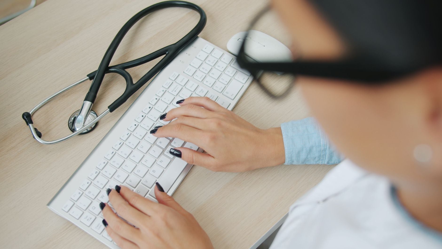 Especialistas revelam o que pode acontecer com o corpo de quem usa computador por mais de 4 horas 1 O médico usa o teclado do computador; o estetoscópio está próximo, sobre a mesa.