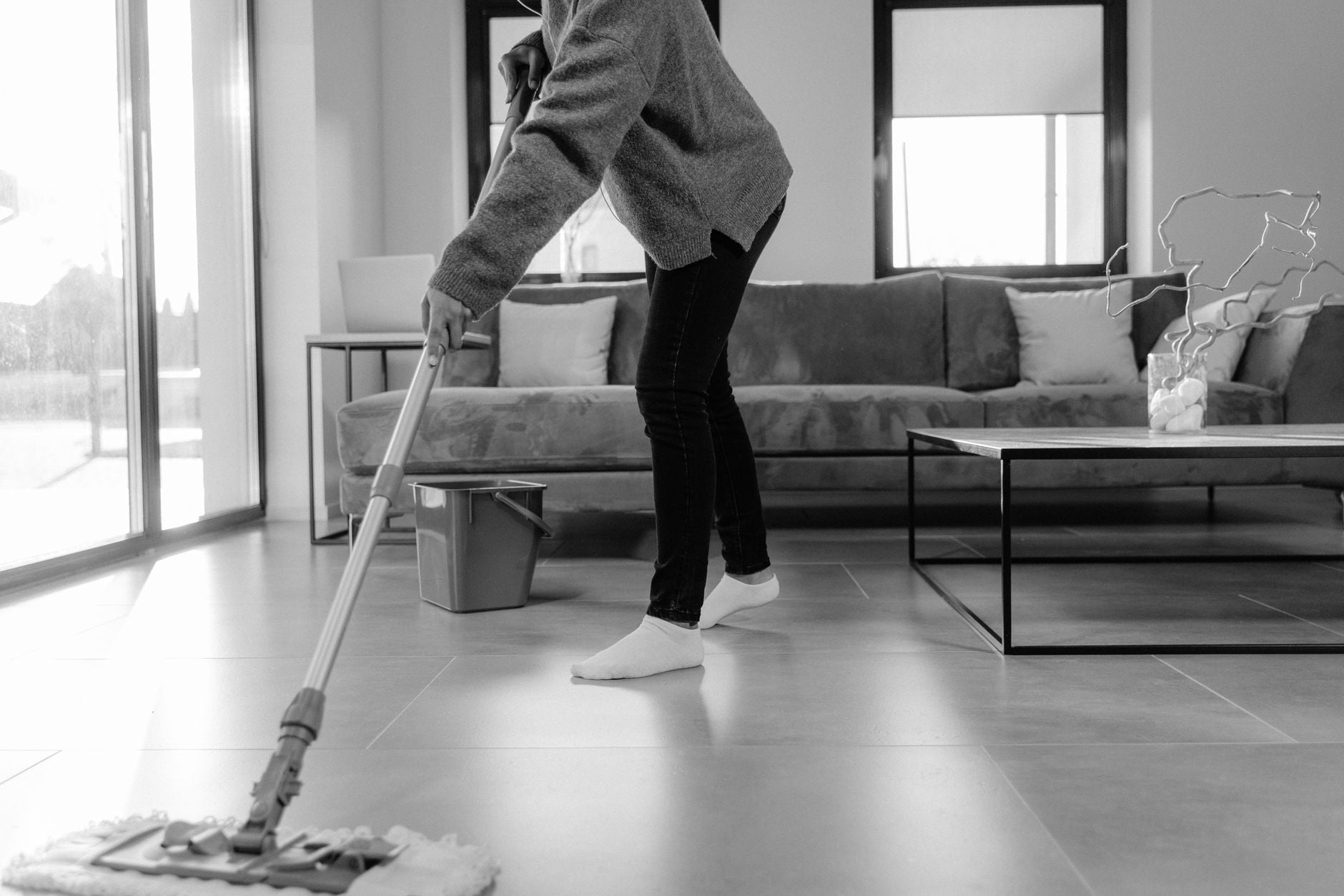 Faxina antes do Natal: a ordem certa para limpar a casa em um dia sem se cansar 1 Uma foto em tons de cinza de uma pessoa limpando o chão em uma sala de estar moderna.
