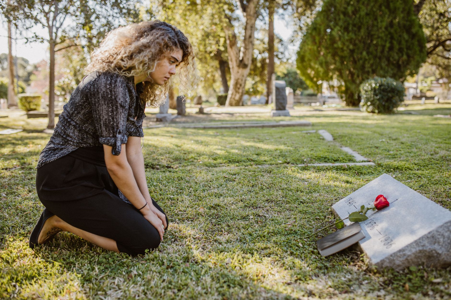 Mesmo sabendo que a morte faz parte da vida, muitas pessoas evitam pensar nela e sentem ansiedade quando o assunto aparece, e a psicologia explica o que realmente está por trás desse medo 1 Uma mulher ajoelhada ao lado de um túmulo em um cemitério iluminado pelo sol, expressando dor e lembrança.