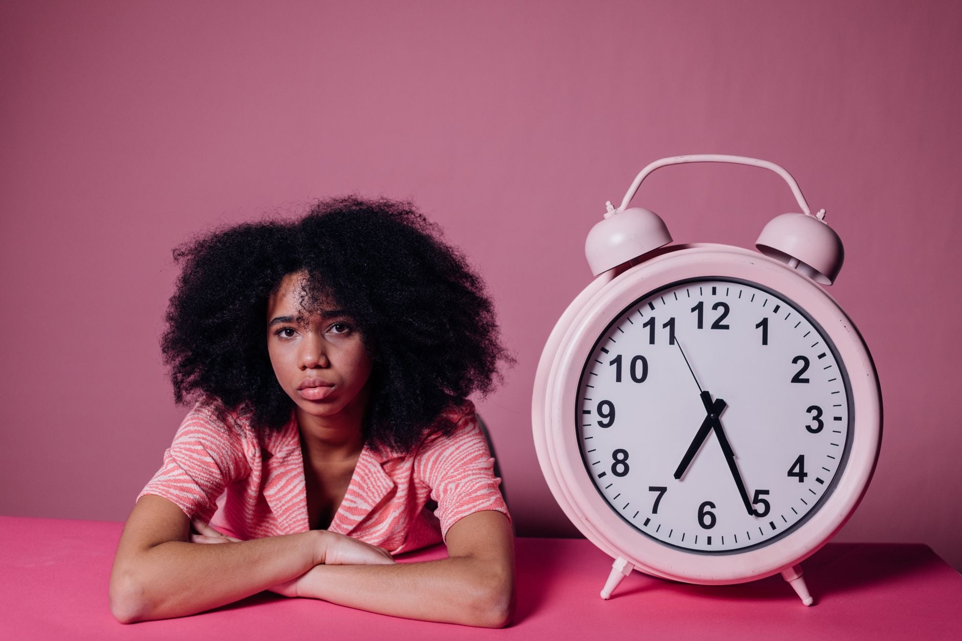 O que a psicologia diz sobre pessoas que deixam tudo para a última hora 2 Uma mulher em uma camisa rosa se apoia em uma mesa ao lado de um grande relógio despertador rosa, com um fundo rosa.