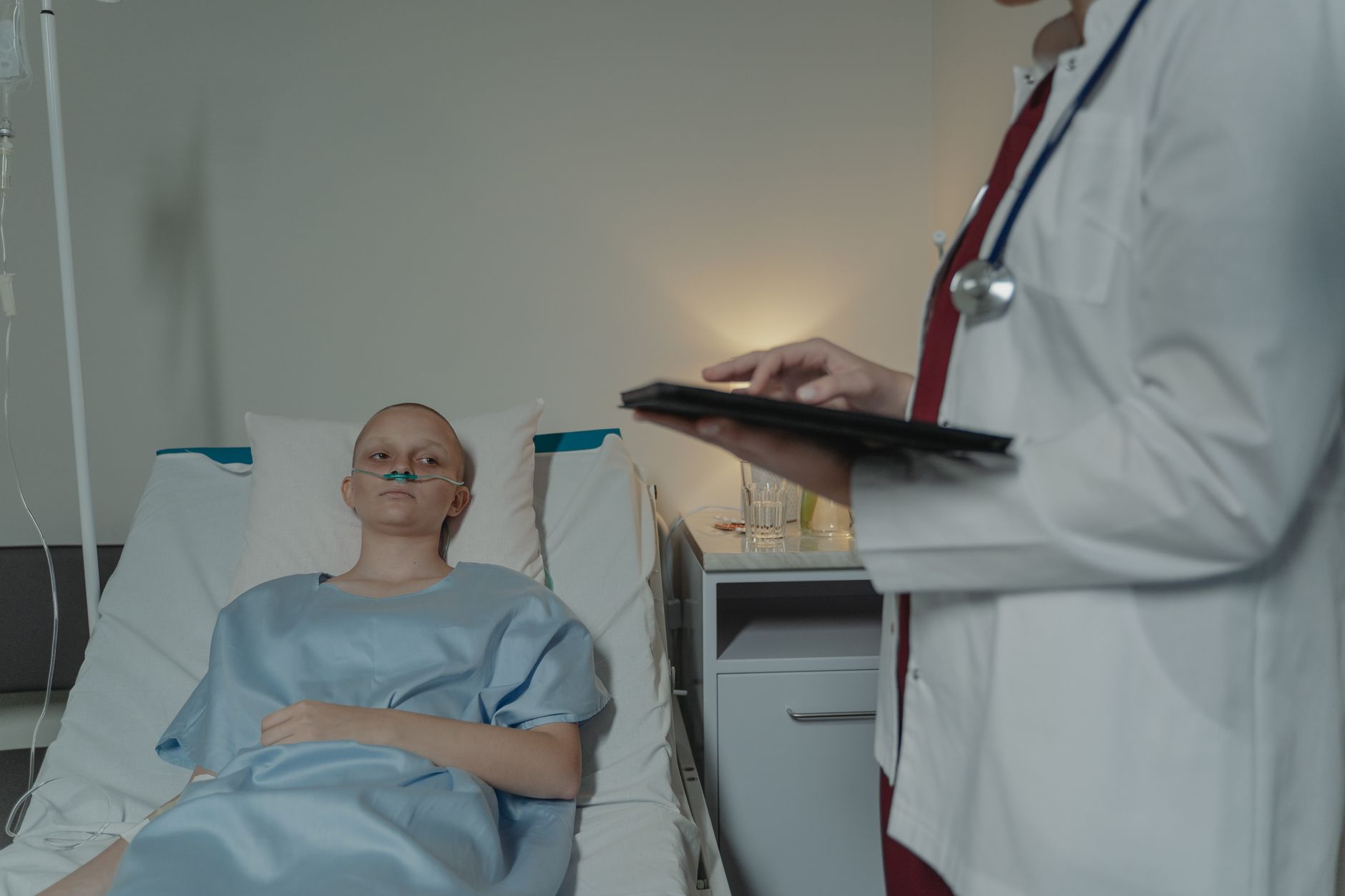 Mulher em uniforme hospitalar deitada na cama, médico verifica tablet digital próximo.