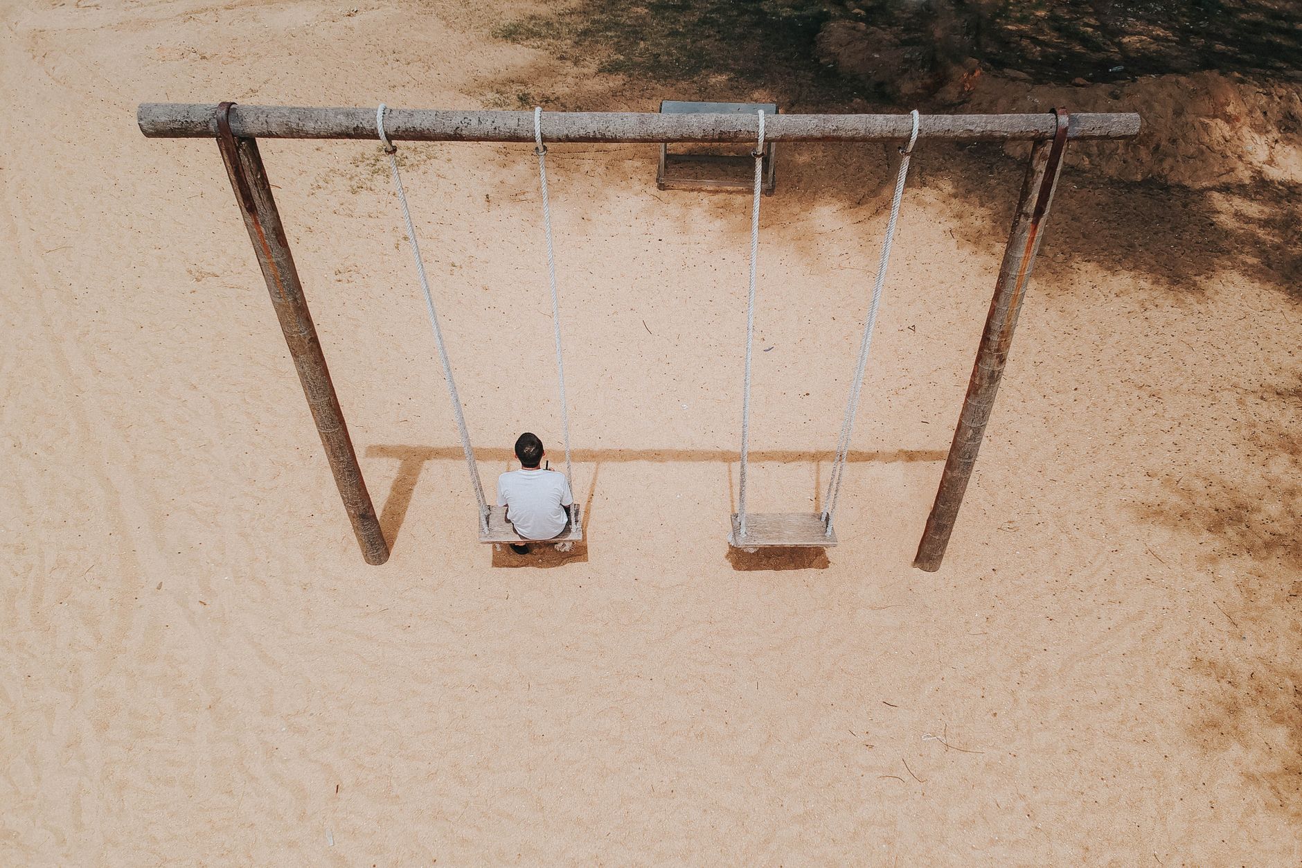 Imagem aérea de um homem sentado sozinho em um balanço em um playground de areia, vista de cima.