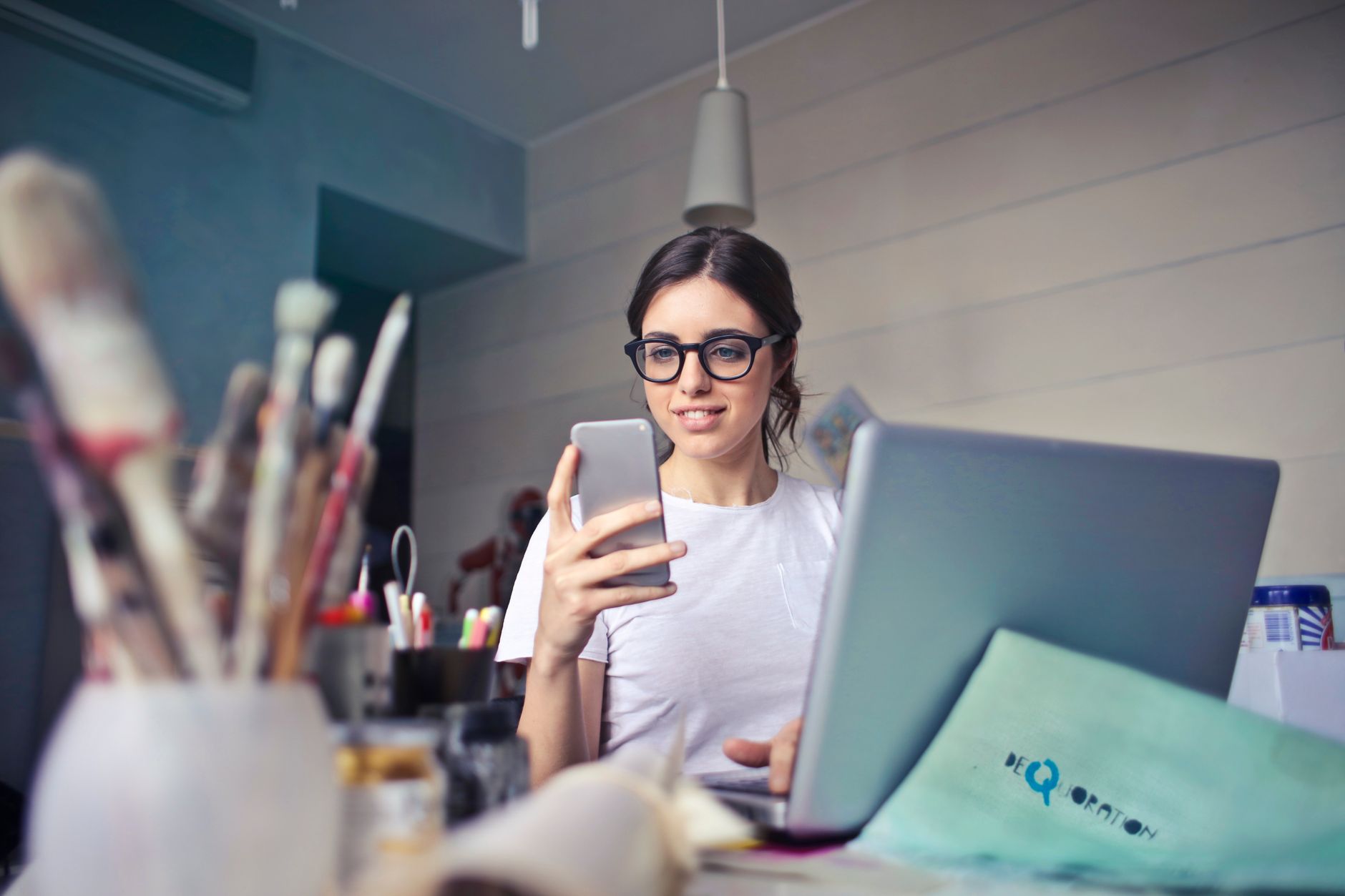 Você conversa sobre um produto e, pouco depois, aparece um anúncio dele no celular; será que os aplicativos estão ouvindo suas conversas? Veja como descobrir 4 Uma mulher usando seu telefone em uma mesa, cercada por materiais de arte e um laptop, em um espaço de trabalho criativo.
