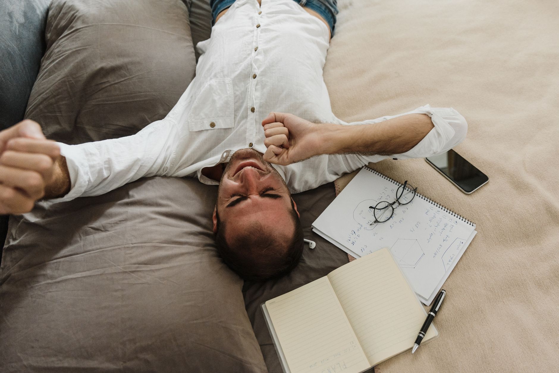 Você costuma dormir depois do almoço e acorda ainda mais cansado? Especialista explica quanto tempo deve durar o cochilo da tarde e por que passar alguns minutos a mais pode atrapalhar sua disposição e o sono da noite 4 Um homem cansado se estica em uma cama cercado por um caderno, caneta e telefone, representando a exaustão do trabalho.